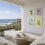 Four framed cocktail art prints with pastel backgrounds hang on a white wall of a modern sunlit patio overlooking the ocean. The prints feature colorful illustrations of cocktails in green, orange, blue, and yellow tones. This image features fine art prints of the Mojito, Strawberry daiquiri, Pina colada and Aperol spritz illustrations by Hannah Singleton at HLSdesigner, inspired by seafood, cuisine and ocean inspired art in Cornwall 