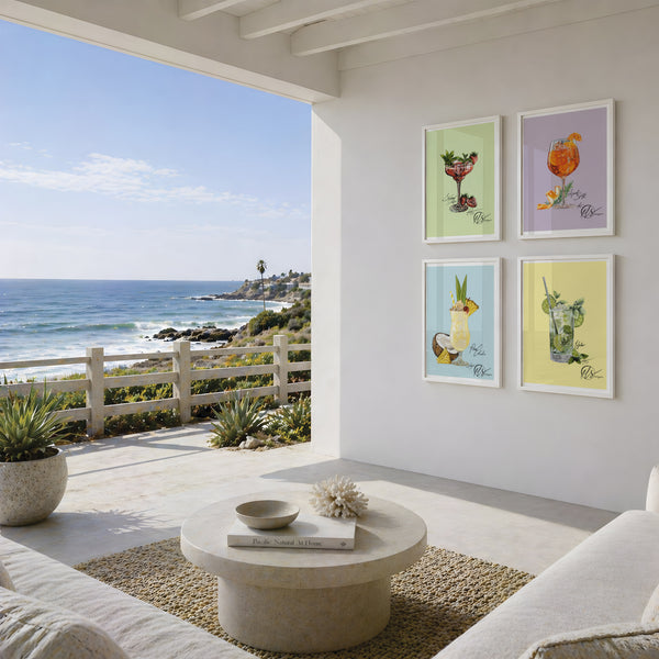 Four framed cocktail art prints with pastel backgrounds hang on a white wall of a modern sunlit patio overlooking the ocean. The prints feature colorful illustrations of cocktails in green, orange, blue, and yellow tones. This image features fine art prints of the Mojito, Strawberry daiquiri, Pina colada and Aperol spritz illustrations by Hannah Singleton at HLSdesigner, inspired by seafood, cuisine and ocean inspired art in Cornwall 