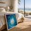 A framed square art print leaning against a bed in a bright, modern bedroom. The artwork depicts a swirling ocean wave in shades of blue and white with black fish silhouettes. Fish shoal 10 - an abstract and expressionist form of colour and textures featuring  bright center with cascading fish swimming around a vortex of ocean as the waves whirl under the surface. Designed by HLSdesigner, Hannah Singleton ocean inspired artist 