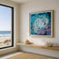 A framed abstract painting featuring a swirling blue, teal, and purple ocean-like pattern with black fish-like shapes, displayed on a beige wall in a modern room with a window overlooking the ocean and a wooden bench below. Fish shoal 11 featured here in a frame on the wall. Featuring lush pinks and lilac sun rays shining through from a unset above the ocean, as a vortex of a water whirlpool steer the sea life below. A shoal of abstract fish swimming amongst the waves rising towards the air above.