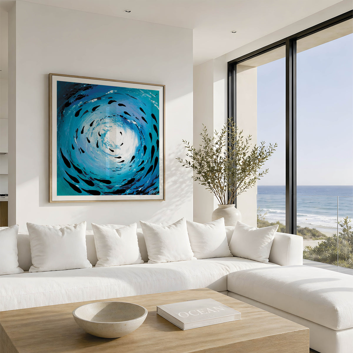 A large framed abstract painting of a swirling blue and black ocean scene hangs on a white wall above a modern white sectional sofa in a bright coastal living room with a wooden coffee table and large windows overlooking the ocean. Fish shoal 12 - through a series of 12 different fish shoal abstract paintings,Hannah has created a beautiful set of prints featuring different textures and colours to for a cohesive set. HLSdesigner ocean inspired art and coastal home decor. Based in Cornwall.