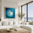 A large framed abstract painting of a swirling blue and black ocean scene hangs on a white wall above a modern white sectional sofa in a bright coastal living room with a wooden coffee table and large windows overlooking the ocean. Fish shoal 12 - through a series of 12 different fish shoal abstract paintings,Hannah has created a beautiful set of prints featuring different textures and colours to for a cohesive set. HLSdesigner ocean inspired art and coastal home decor. Based in Cornwall.