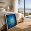 A framed square art print leaning against a bed in a bright beach-view bedroom. The artwork shows a swirling blue underwater scene with black fish. Fish shoal 2 - image not to scale - showcasing the fine art print by HLSdesigner, Cornwall based artist Hannah Singleton. 
