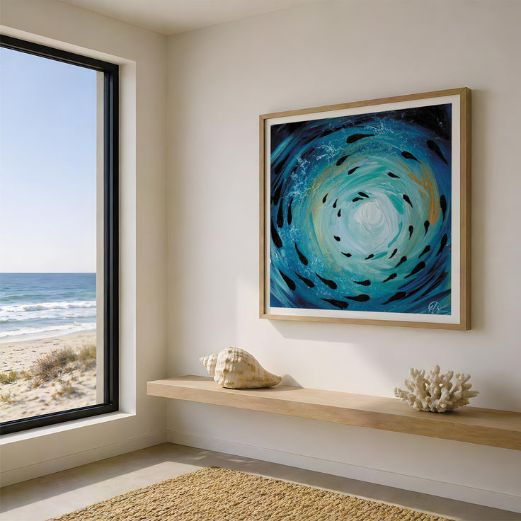 A framed square wall art print depicting a swirling blue and turquoise ocean scene with black fish, displayed in a modern room with a window overlooking the beach and ocean. Fish shoal 3 - depicting an abstract and expressionist form of an ocean vortex with fish swimming throughout the motion of the sea, with gold hues and bright light reflecting through the painting to show sunlight reflecting through the surface. A coastal inspired underwater themed art print designed by Hannah Singleton at HLSdesigner