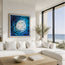 A framed abstract blue and white swirl painting hangs on a white wall above a modern white sectional sofa in a bright coastal living room with large windows overlooking the ocean. Fish shoal 5 - not to scale - fish swimming around under the ocean as the sun reflects through the surface, painted in an abstract form. designed by Hannah singleton at HLSdesigner, ocean inspired cornwall based artist 