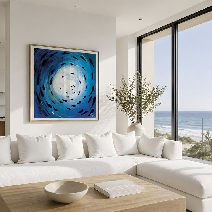 A framed abstract blue and white swirl painting hangs on a white wall above a modern white sectional sofa in a bright coastal living room with large windows overlooking the ocean. Fish shoal 5 - not to scale - fish swimming around under the ocean as the sun reflects through the surface, painted in an abstract form. designed by Hannah singleton at HLSdesigner, ocean inspired cornwall based artist 