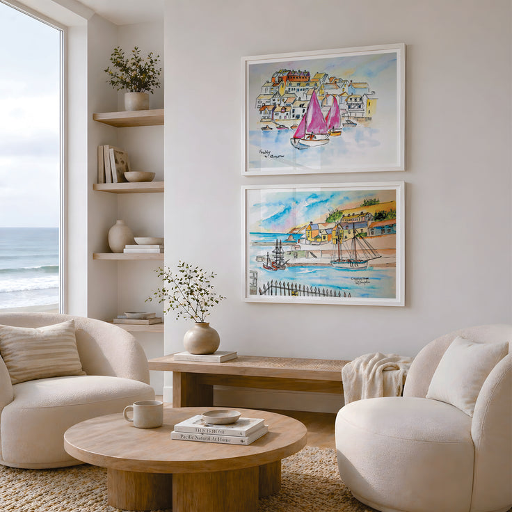 Two framed watercolour art prints depicting coastal scenes and sailboats, displayed on a light beige wall in a modern, sunlit living room with cream armchairs and a round wooden coffee table. These art prints are reproductions of paintings by Robert Singleton featuring Fowey and the racing yachts and Charlestown with the tall ships. Modern, contemporary and colourful, inspired by the coastal towns of Cornwall