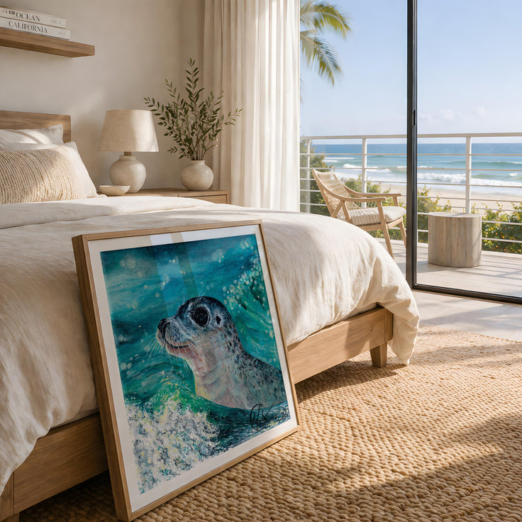 A close up image featuring gentle curiosity - a fine art print of a seal pup popping its head out of the waves in a summery glistening ocean. Bold innocent eyes and pretty facial features, this image displays the print in a wooden frame and lies against the end of a bed in a coastal beach house colour scheme. Image is for representation only and does not show the scale of the print accurately, please see size details on your selection before purchase. Designed and made by HLSdesigner, Hannah Singleton