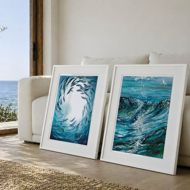 Two framed blue ocean art prints leaning against a white sofa in a bright living room, with a window showing the sea and sky in the background. This is shoal under surface and glistening waters fine art prints created by HLSdesigner - Hannah Singleton ocean inspired fine artist and illustrator from Cornwall. Inspired by unique viewpoints of the ocean, underwater and waves. focusing on reflections on water. These prints feature cool blue and green tones with a pop of colour added in. Two beautiful works