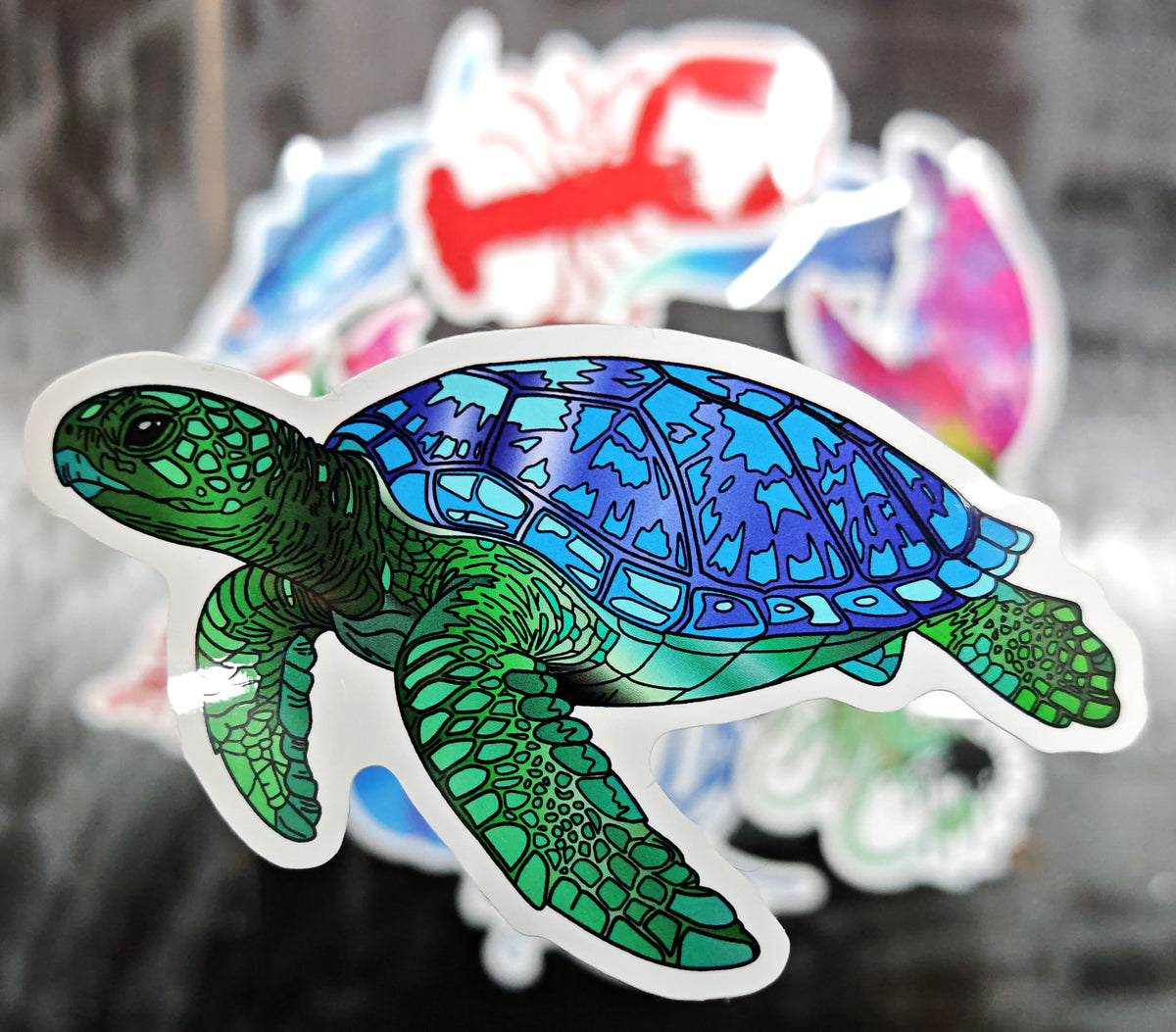 green and blue sea turtle sticker raised in the foreground with various stickers in the background blurred 