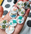 Hand holding various small round pin badges with different designs on a patterned surface.