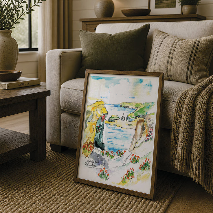 A framed watercolor landscape painting leaning against a beige sofa in a cozy living room setting. The painting depicts a scenic coastal landscape of the picturesque Cornish destination that is Kynance cove. Using minimal watercolour and linear strokes, Robert Singleton, contemporary artist created this stunning piece. Perfect for minimalistic wall space or a cozy home area. This painting creates a pop of colour anywhere. Reproduced into fine art prints exclusively by HLSdesigner