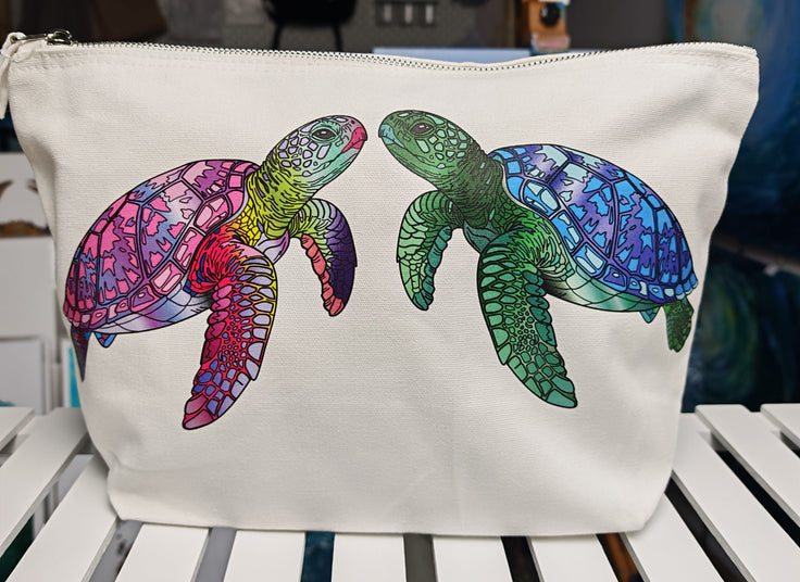travel accessory bag for makeup or cosmetics and gadgets, in off white colour, showing two turtles facing each other. one pink and purple and yellow and the other green and blue, complete with a silver zip and holing 5 litre capacity. bag is sitting on a white shelf with ocean art in the background