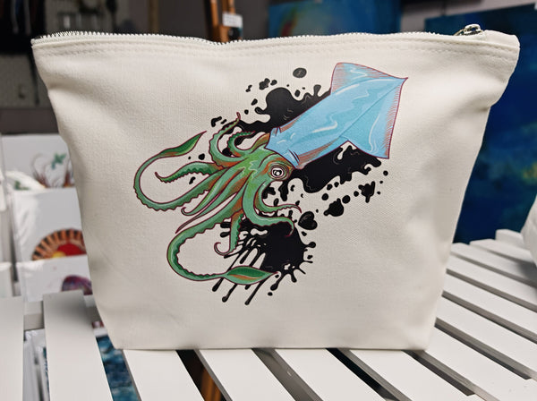 squid ink large cosmetic bags, illustration of a squid positioned diagonal with ink splatters behind it and black ink hearts around it, in blue with green and orange tentacles pressed onto an off white cosmetic bag with a silver zip and fabric pull handle.  this is in the large size and is sitting on a white shelf with ocean artworks behind it in the background, capacity of this bag is 5 litres