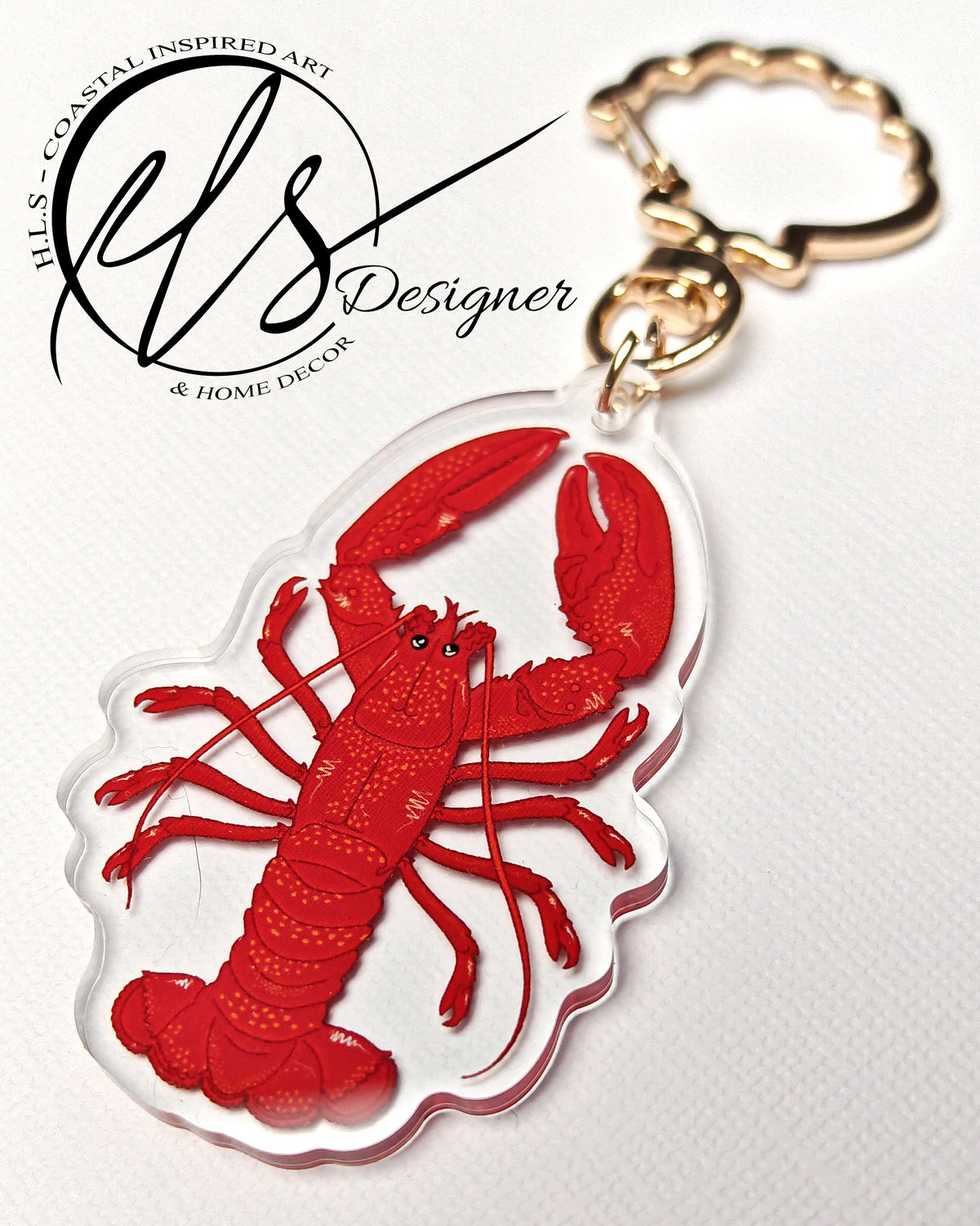 Keychain made from clear acrylic with red lobster design with orange spots and white details on a white background, finished with a seashell shaped metal keyring clasp featuring a logo in the corner reading, HLSdesigner HLS coastal inspired art and home décor 