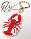 Keychain made from clear acrylic with red lobster design with orange spots and white details on a white background, finished with a seashell shaped metal keyring clasp featuring a logo in the corner reading, HLSdesigner HLS coastal inspired art and home décor 