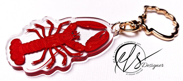 Red lobster-shaped keychain with a clear background, close up photo showing the depth of the acrylic keychain. clear acrylic with a red and orange lobster illustration finished with a gold metal seashell clasp with the HLSdesigner logo 