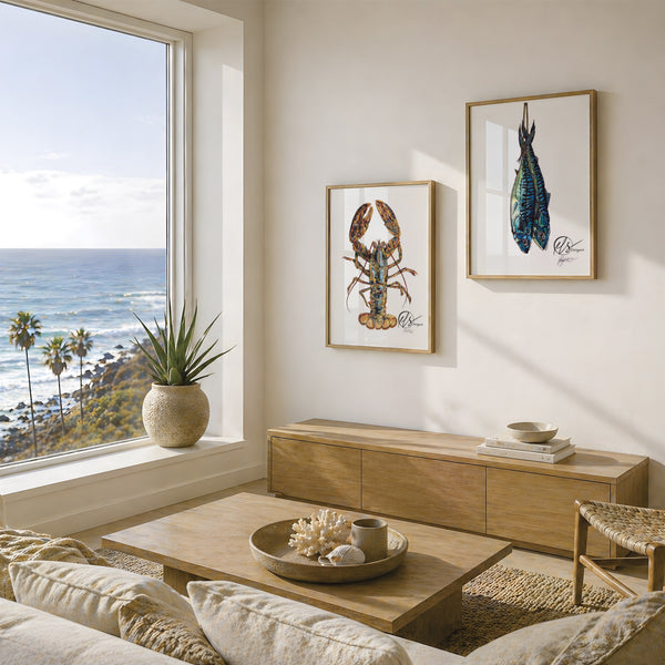 A styled living room corner with two framed lobster and fish art prints on a cream wall above a light wood media console, with a large window showing a beach and ocean view. This is a pair of fine art prints by HLSdesigner featuring a realistic illustration of a lobster on a white background and another of two mackerel hanging after being caught, beautiful prints for kitchen or nautical home décor. 