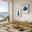 A styled living room interior with two framed blue fish art prints hanging on a cream wall above a wooden media console. A large window shows the ocean and palm trees, with a sofa, coffee table, and potted plant visible in the room. Fine art prints feature two mackerel fish roped after catch ready for market and a handful of mussels from the seafood platter range. Illustrated by Hannah Singleton at HLSdesigner, coastal fine artist in Cornwall