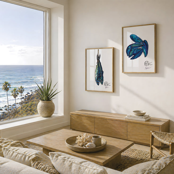 A styled living room interior with two framed blue fish art prints hanging on a cream wall above a wooden media console. A large window shows the ocean and palm trees, with a sofa, coffee table, and potted plant visible in the room. Fine art prints feature two mackerel fish roped after catch ready for market and a handful of mussels from the seafood platter range. Illustrated by Hannah Singleton at HLSdesigner, coastal fine artist in Cornwall
