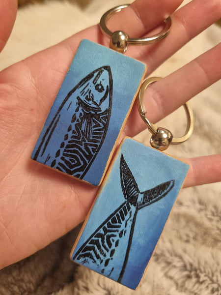 two hand painted wooden keyrings with mackerel designs. one has a mackerel head and the other the mackerel tail , both in an Aztec style in black on a blue background. a hand holds them up to the camera over a fur backing