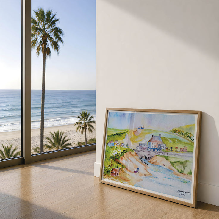 A framed landscape watercolour painting leaning against a white wall on a light wood floor, showing a beach, ocean, palm trees, and a coastal village in the art print - this is the village of Mawgan Porth overlooking the beach, a contemporary and modern approach to showcase the beauty in Cornwall. The painting is displayed in a modern room setting with a large window. Originally painted by Robert Singleton - this has been lovingly reproduced into fine art prints exclusively by HLSdesigner