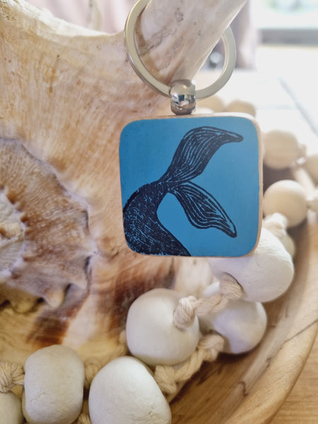 Square blue pendant keyring with a black mermaid tail design on a shell and stones background
