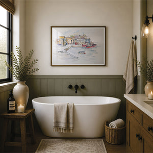 A framed watercolor painting of a harbor scene hangs on a beige wall above a white freestanding bathtub in a styled bathroom with wood wainscoting, a window, candles, towels, and a vanity. The picture in the frame is a reproduction print of an original watercolour painting by Robert singleton and produced exclusively by HLSdesigner - this is a print featuring mevagissey - a beautiful coastal town in the heart of Cornwall 
