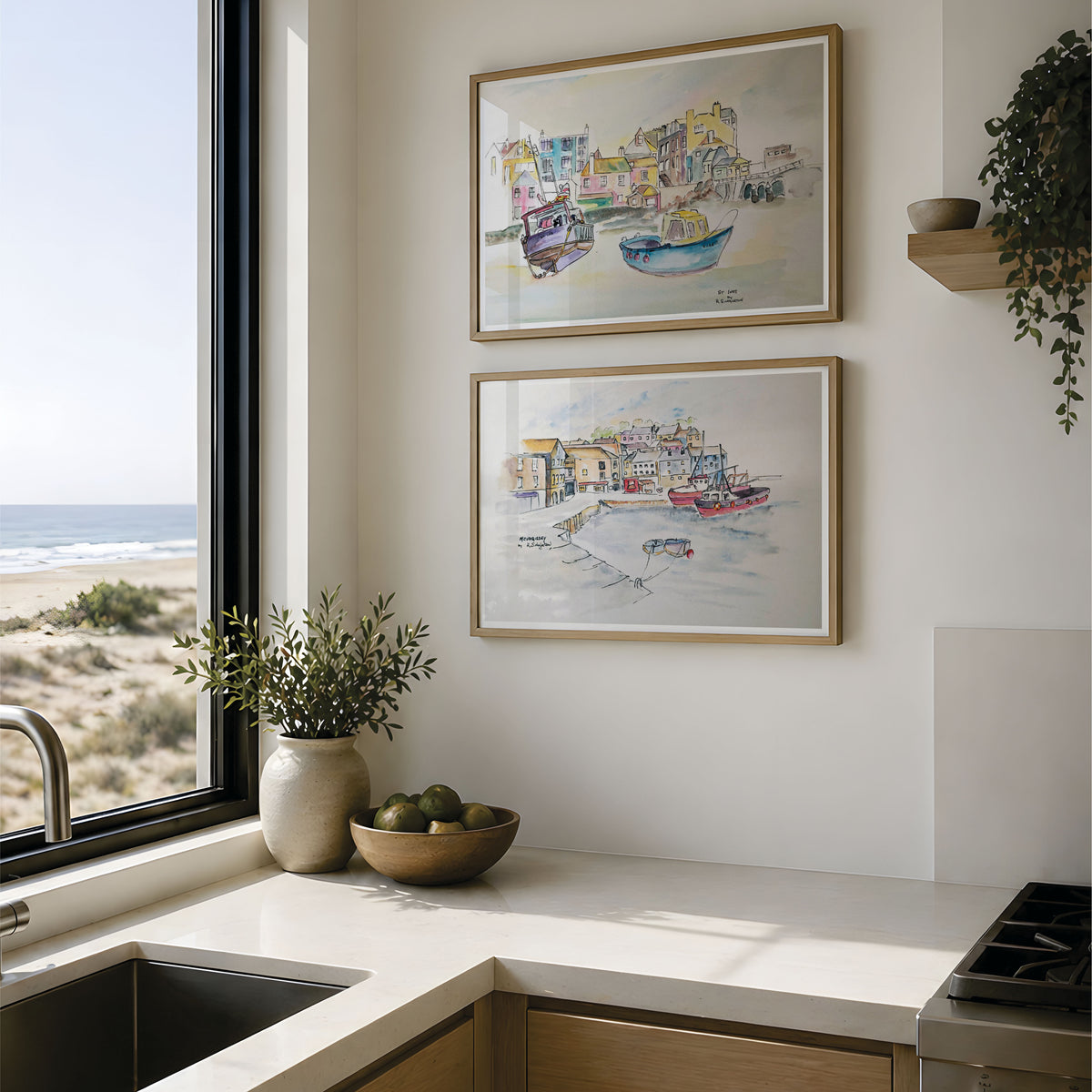 Two framed watercolor paintings of coastal harbor scenes with boats and colorful buildings, displayed on a white wall above a kitchen counter with a window view of the beach and ocean. This pair of fine art pints are reproductions from Robert singleton and his contemporary original watercolour studies of the towns around cornwall. Mevagissey and st ives are pictured in this mock up. 