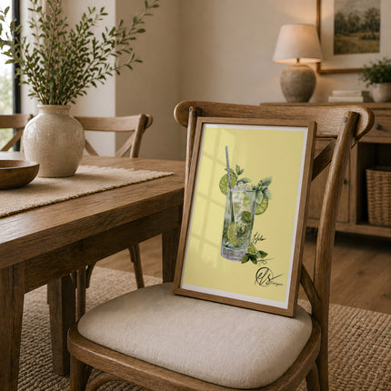 A framed art print featuring an illustration of a mojito cocktail on a pale yellow background, leaning on a wooden dining chair in a warm, modern dining room setting. HLSdesigner has illustrated a glass filled with a Mojito with limes and mint leaves to create a realistic yet stylised portrayal of a beautiful summer cocktail. Inspired by foodies and beach days in cornwall.