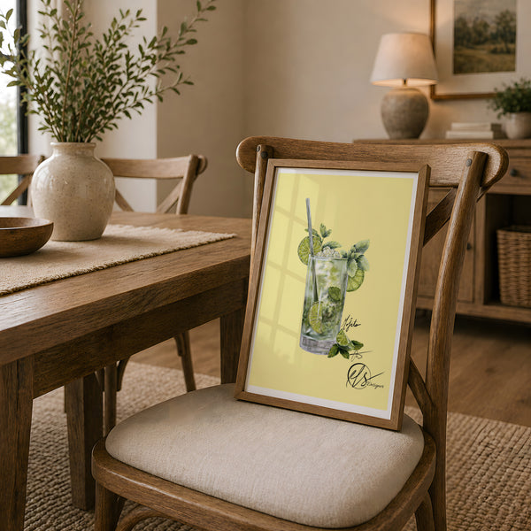 A framed art print featuring an illustration of a mojito cocktail on a pale yellow background, leaning on a wooden dining chair in a warm, modern dining room setting. HLSdesigner has illustrated a glass filled with a Mojito with limes and mint leaves to create a realistic yet stylised portrayal of a beautiful summer cocktail. Inspired by foodies and beach days in cornwall.