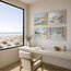 Four framed coastal landscape art prints are displayed in a grid on a beige wall above a white desk and chair in a bright, modern room with a large window overlooking a sandy beach and ocean. These are reproduction art prints of Robert Singleton - contemporary and minimalist watercolour paintings of Sennen cove, Porth beach, Perranporth beach and Newquay harbour - inspired by the coastal towns of Cornwall. Reproduced exclusively by HLSdesigner