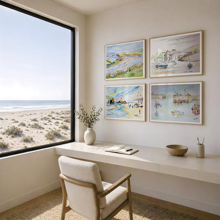 Four framed coastal landscape art prints are displayed in a grid on a beige wall above a white desk and chair in a bright, modern room with a large window overlooking a sandy beach and ocean. These are reproduction art prints of Robert Singleton - contemporary and minimalist watercolour paintings of Sennen cove, Porth beach, Perranporth beach and Newquay harbour - inspired by the coastal towns of Cornwall. Reproduced exclusively by HLSdesigner