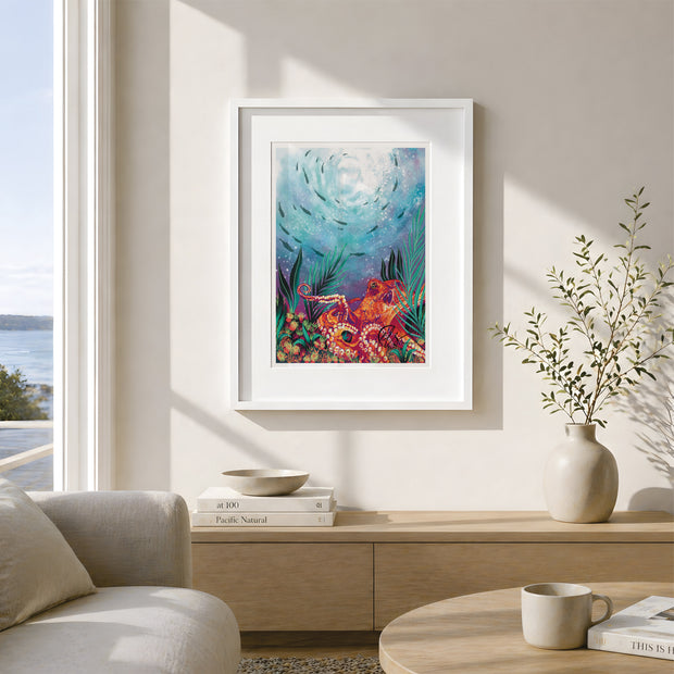 A framed art print featuring a colourful underwater scene with sea life, displayed on a beige wall in a modern, sunlit living room setting. Featuring a bright orange octopus on a seabed of coral with seaweed and a shoal of fish swimming in the background under the reflections of the sun from the surface of the sea. Designed and created by Hannah Singleton fine artist and illustrator in Cornwall inspired by underwater and surface viewpoints of the sea and the Cornish coast. HLSdesigner