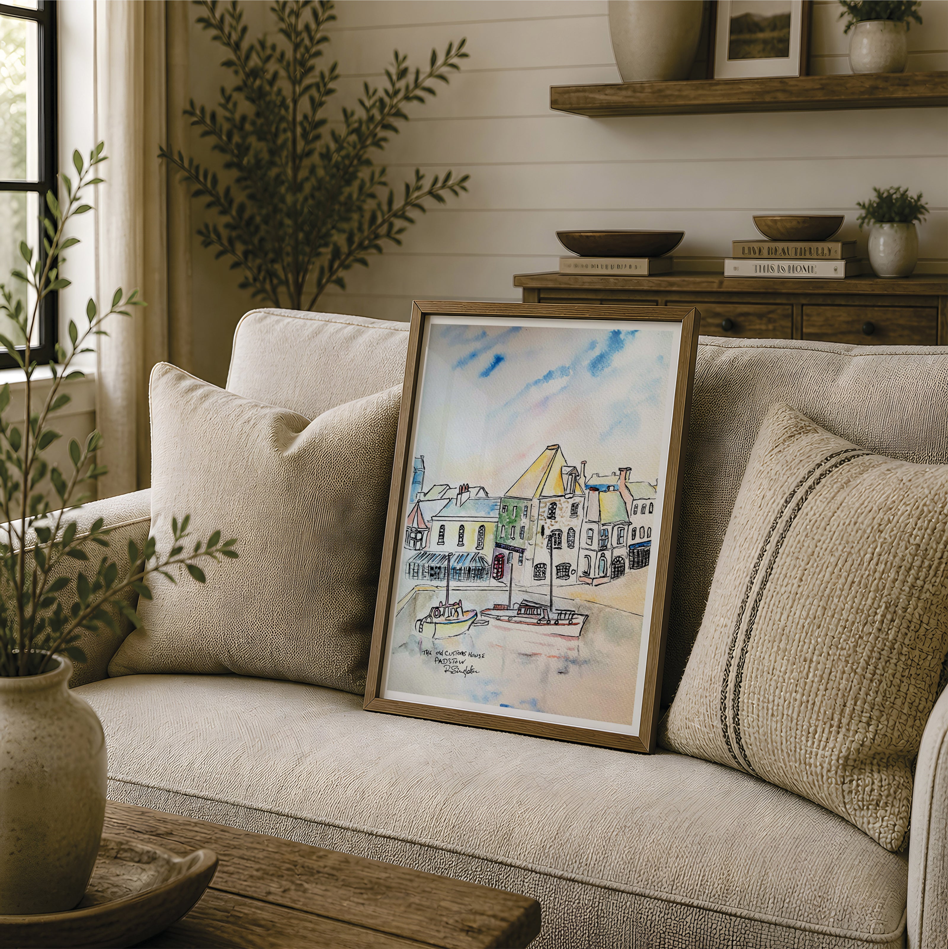 A framed watercolour painting of a coastal harbour town with boats, leaning against a beige sofa in a styled living room setting. This painting focuses on the old customs house - a famous house in Cornwall with fishing boats in the harbour below. Reproduced exclusively by HLSdesigner in tribute of Robert Singleton