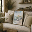 A framed watercolour painting of a coastal harbour town with boats, leaning against a beige sofa in a styled living room setting. This painting focuses on the old customs house - a famous house in Cornwall with fishing boats in the harbour below. Reproduced exclusively by HLSdesigner in tribute of Robert Singleton