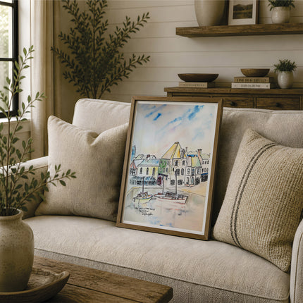 A framed watercolour painting of a coastal harbour town with boats, leaning against a beige sofa in a styled living room setting. This painting focuses on the old customs house - a famous house in Cornwall with fishing boats in the harbour below. Reproduced exclusively by HLSdesigner in tribute of Robert Singleton