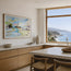 A framed coastal landscape painting hangs on a light beige wall in a modern kitchen with wood cabinetry, a stone countertop, and a large window overlooking the ocean. This is Perranporth beach. A watercolour study depicting the essence of what is perranporth and the beach goers enjoying it. A coastal seascape beach scene originally painted in watercolour by artist Robert singleton showcasing his minimalist contemporary work. Reproduced exclusively by Hannah singleton at HLSdesigner.