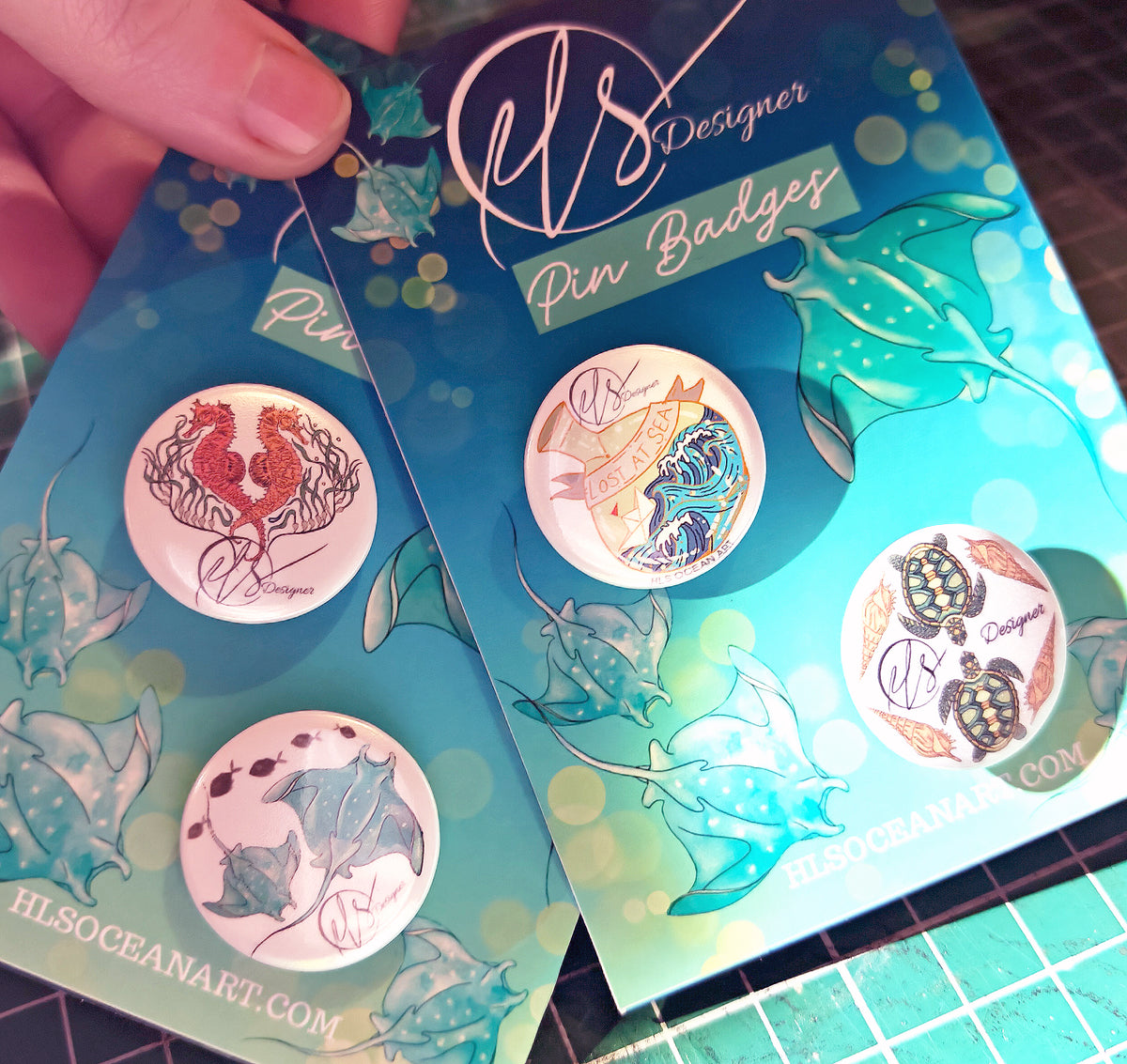 Two packs of pin badges with ocean-themed designs on a cutting mat.