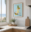 A framed tropical cocktail art print hangs on a white kitchen wall above a countertop with a vase of greenery and a wooden cutting board. The print shows a pina colada cocktail drink with pineapple, a coconut half, and light blue background. Designed by Hannah Singleton at HLSdesigner, colourful cocktail fine art prints and ocean paintings for a nautical beach inspired home décor range. made in Cornwall 