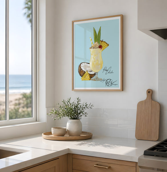 A framed tropical cocktail art print hangs on a white kitchen wall above a countertop with a vase of greenery and a wooden cutting board. The print shows a pina colada cocktail drink with pineapple, a coconut half, and light blue background. Designed by Hannah Singleton at HLSdesigner, colourful cocktail fine art prints and ocean paintings for a nautical beach inspired home décor range. made in Cornwall 