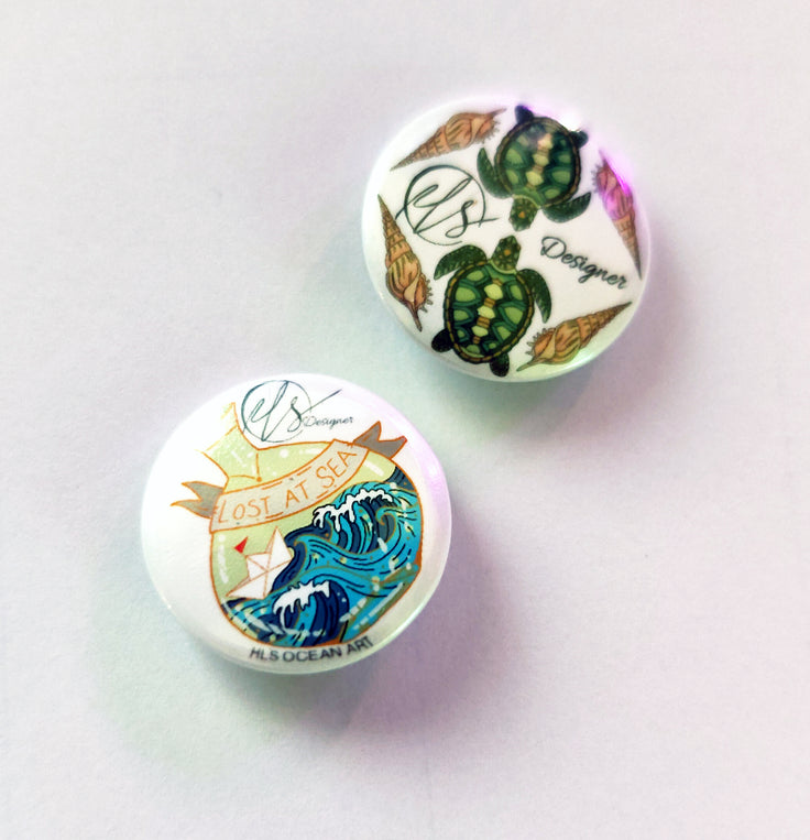 two circular pin badges collectable as a set of two, one with a message in a bottle design on and one with turtles and shells with the HLSdesigner logo, designed and made in house