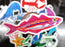 raised in the foreground with blurred stickers in the background on a dark surface, pink sardine die cut vinyl sticker