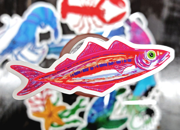 raised in the foreground with blurred stickers in the background on a dark surface, pink sardine die cut vinyl sticker