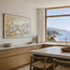 A framed watercolor painting of a coastal village hangs on a light beige wall in a modern kitchen with a large window overlooking the blue ocean and rocky coastline. Polperro in Colour, a reproduction fine art print of a contemporary watercolour painting by Robert Singleton - focusing on the beauty of the smal Cornish town of Polperro featuring the fishing village buildings in a colourful minimal style