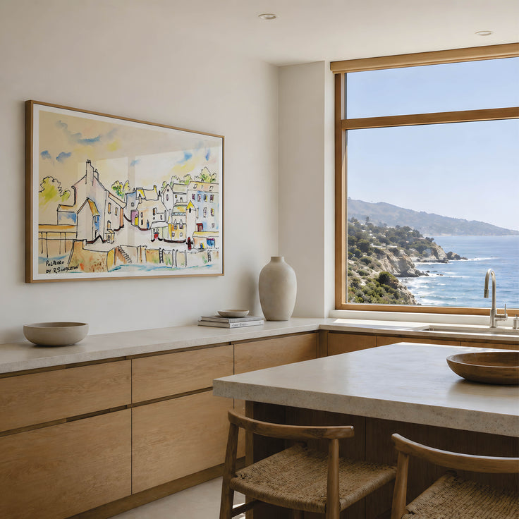 A framed watercolor painting of a coastal village hangs on a light beige wall in a modern kitchen with a large window overlooking the blue ocean and rocky coastline. Polperro in Colour, a reproduction fine art print of a contemporary watercolour painting by Robert Singleton - focusing on the beauty of the smal Cornish town of Polperro featuring the fishing village buildings in a colourful minimal style