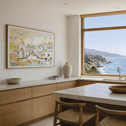 A framed watercolor painting of a coastal village hangs on a light beige wall in a modern kitchen with a large window overlooking the blue ocean and rocky coastline. Polperro in Colour, a reproduction fine art print of a contemporary watercolour painting by Robert Singleton - focusing on the beauty of the smal Cornish town of Polperro featuring the fishing village buildings in a colourful minimal style