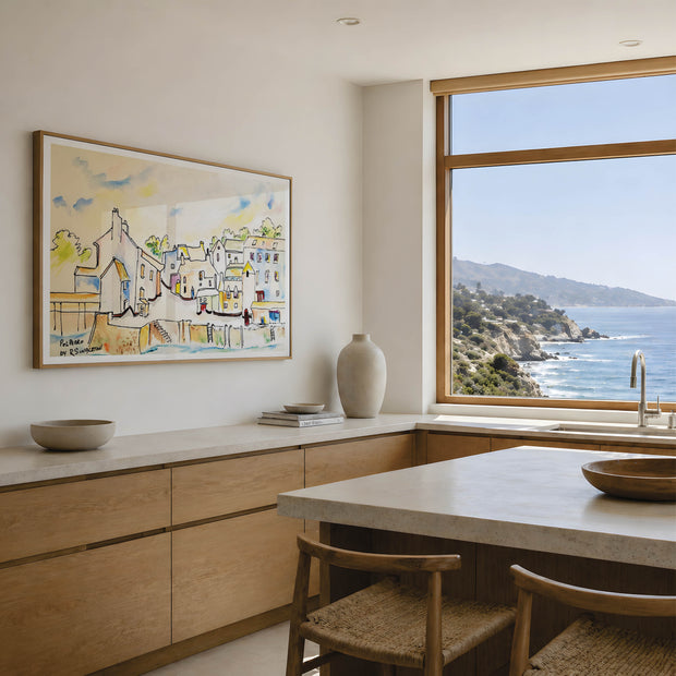 A framed watercolor painting of a coastal village hangs on a light beige wall in a modern kitchen with a large window overlooking the blue ocean and rocky coastline. Polperro in Colour, a reproduction fine art print of a contemporary watercolour painting by Robert Singleton - focusing on the beauty of the smal Cornish town of Polperro featuring the fishing village buildings in a colourful minimal style