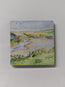 a square wooden fridge magnet with a printed watercolour painting of Porth beach in Newquay Cornwall. painted using colour washes and use of negative space by R Singleton - tribute collection reproduced exclusively by HLSdesigner, showing the beach from the higher footpath where a bench lies vacant to take in the views of the rolling in stream and the camp site behind. 