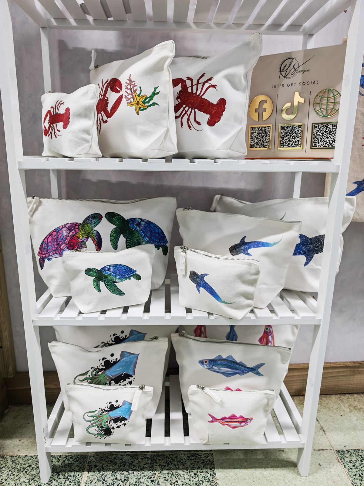 designed and printed in house at hlsdesigner, view the full range of cosmetic accessory bags in all sizes, small, medium and large. set on a white shelving unit complete with a QR coded social media poster directing to facebook, tiktok and online. on the top shelf is a lobster collection complete with a starfish and seaweed. On the middle shelf is the turtle collection and the whale shark collection, and on the bottom shelf is an inky squid collection and a colourful pink and blue sardine collection.